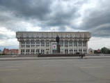 A large, rectangular government-style building with multiple windows and flags on top. In front of the building stands a tall statue on a pedestal. The scene is set against a backdrop of overcast skies, and there are a few people visible on the street near the building.