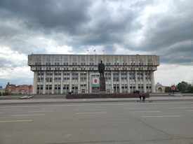 A large, rectangular government-style building with multiple windows and flags on top. In front of the building stands a tall statue on a pedestal. The scene is set against a backdrop of overcast skies, and there are a few people visible on the street near the building.