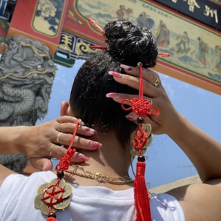 A person with neatly styled hair is adorned with vibrant red and gold traditional Chinese decorations. The hands show manicured nails with red and gold designs. A decorative archway with intricate carvings and Chinese characters is visible in the background.