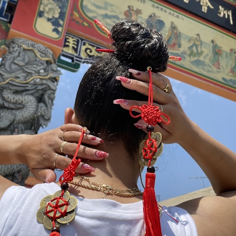 A person with neatly styled hair is adorned with vibrant red and gold traditional Chinese decorations. The hands show manicured nails with red and gold designs. A decorative archway with intricate carvings and Chinese characters is visible in the background.