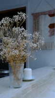 A glass vase filled with delicate white flowers on long, thin stems is placed on a light marble surface. Next to it is a white candle on a small plate. In the background, a woven wall hanging with geometric patterns in muted tones adds a cozy touch.