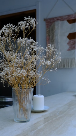 A glass vase filled with delicate white flowers on long, thin stems is placed on a light marble surface. Next to it is a white candle on a small plate. In the background, a woven wall hanging with geometric patterns in muted tones adds a cozy touch.