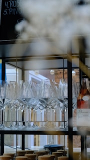 A shelf displays an arrangement of neatly organized wine glasses, with a bottle of wine situated on the right. The background includes a blurred view of a menu board with handwritten text. The setting appears to be a bar or a restaurant with soft lighting.