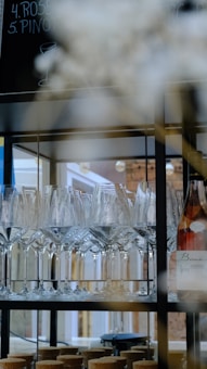 A shelf displays an arrangement of neatly organized wine glasses, with a bottle of wine situated on the right. The background includes a blurred view of a menu board with handwritten text. The setting appears to be a bar or a restaurant with soft lighting.