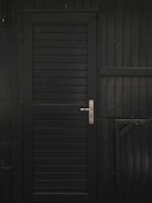 A vertically slatted black door with a metallic handle, set in a matching dark wooden wall with horizontal slats and reinforced beams.
