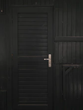 A vertically slatted black door with a metallic handle, set in a matching dark wooden wall with horizontal slats and reinforced beams.