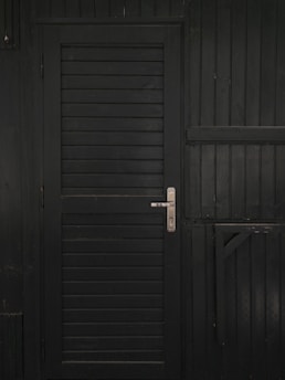 A vertically slatted black door with a metallic handle, set in a matching dark wooden wall with horizontal slats and reinforced beams.