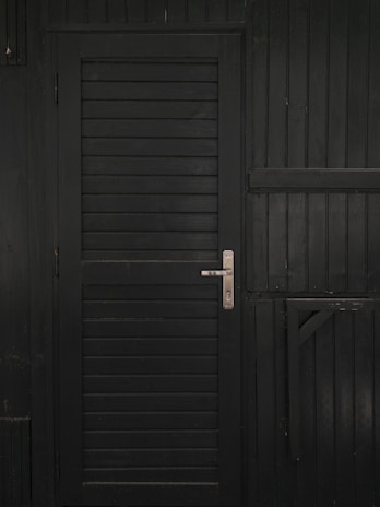 A vertically slatted black door with a metallic handle, set in a matching dark wooden wall with horizontal slats and reinforced beams.