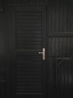 A vertically slatted black door with a metallic handle, set in a matching dark wooden wall with horizontal slats and reinforced beams.