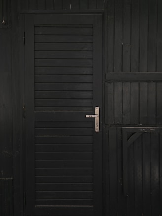 A vertically slatted black door with a metallic handle, set in a matching dark wooden wall with horizontal slats and reinforced beams.