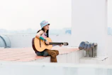 A scenic outdoor shot of Hailey sitting on a rooftop at sunset, guitar in hand.