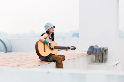 A scenic outdoor shot of Hailey sitting on a rooftop at sunset, guitar in hand.