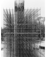 An intricate lattice of steel rebar forms a complex grid structure on a construction site. The metallic framework is part of the initial phase of a concrete structure's reinforcement, standing vertically with a highly geometric and organized pattern. The background includes some natural elements like trees and a clear sky.