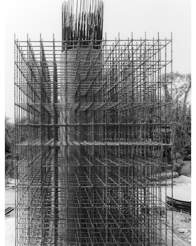 An intricate lattice of steel rebar forms a complex grid structure on a construction site. The metallic framework is part of the initial phase of a concrete structure's reinforcement, standing vertically with a highly geometric and organized pattern. The background includes some natural elements like trees and a clear sky.