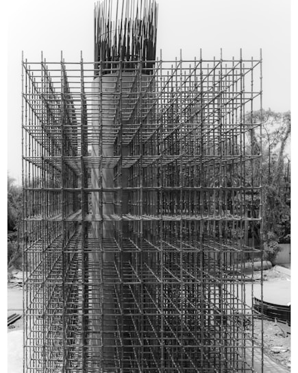 An intricate lattice of steel rebar forms a complex grid structure on a construction site. The metallic framework is part of the initial phase of a concrete structure's reinforcement, standing vertically with a highly geometric and organized pattern. The background includes some natural elements like trees and a clear sky.