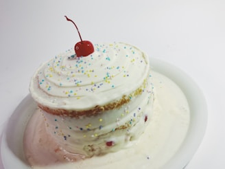 A layered cake with white frosting, topped with a cherry and sprinkled with multicolored round sprinkles. The cake is placed on a white plate, and some of the frosting appears to be melting and pooling at the base.
