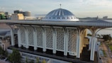A modern architectural structure with a large, metallic dome roof and intricate geometric patterns on the facade. The building is surrounded by urban development, with water features and trees nearby. The sky is partially cloudy, suggesting a calm weather condition.