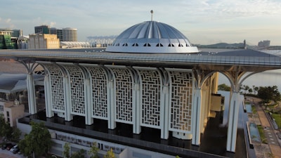 A modern architectural structure with a large, metallic dome roof and intricate geometric patterns on the facade. The building is surrounded by urban development, with water features and trees nearby. The sky is partially cloudy, suggesting a calm weather condition.