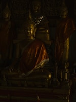 A grouping of intricately designed Buddha statues, each adorned with ornate golden robes. The ambient lighting casts deep shadows, highlighting the serene expressions of the statues. Smaller figures are arranged at the base, adding to the overall spiritual ambiance.