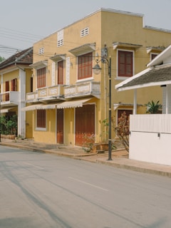 A row of charming, two-story colonial-style buildings with yellow facades and brown wooden shutters. Decorative white trim accents the windows and roof edges. The street is quiet and lined with potted plants and a single streetlamp. The scene is well-lit with natural daylight, casting soft shadows.