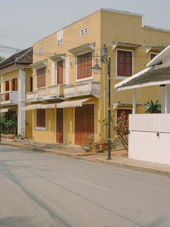 A row of charming, two-story colonial-style buildings with yellow facades and brown wooden shutters. Decorative white trim accents the windows and roof edges. The street is quiet and lined with potted plants and a single streetlamp. The scene is well-lit with natural daylight, casting soft shadows.