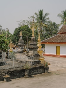 The image features a traditional temple courtyard with ornate stone structures topped with spires. Several tropical trees, including palm trees, are visible in the background. A building with white walls and a brown, pitched roof is on the right side. The area is surrounded by greenery and appears to be calm and serene.