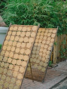 Two bamboo drying racks are positioned outdoors, each covered with round pieces of flatbread or rice crackers aligned in a grid pattern. Behind the racks, lush green bamboo plants create a natural backdrop. The ground is made of brick paving, adding a rustic atmosphere to the scene.