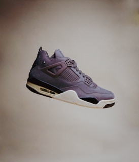 A single sneaker with a sleek, modern design is visibly suspended in mid-air against a plain background. The shoe features a mix of materials, including smooth leather and mesh, with intricate detailing around the lace area and a distinctive air cushion in the sole.