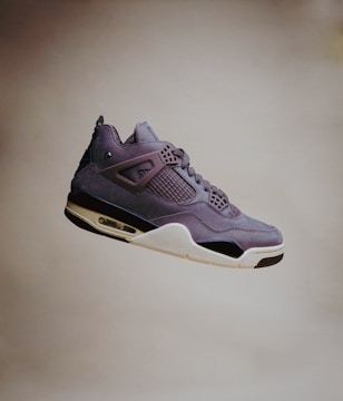 A single sneaker with a sleek, modern design is visibly suspended in mid-air against a plain background. The shoe features a mix of materials, including smooth leather and mesh, with intricate detailing around the lace area and a distinctive air cushion in the sole.