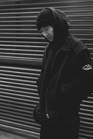 A person dressed in dark clothing, including a hood and beanie, stands in front of a corrugated metal background. The monochrome image has a strong contrast, enhancing the shadows and giving a dramatic appearance.