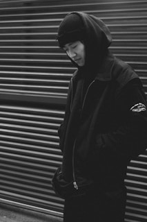 A person dressed in dark clothing, including a hood and beanie, stands in front of a corrugated metal background. The monochrome image has a strong contrast, enhancing the shadows and giving a dramatic appearance.