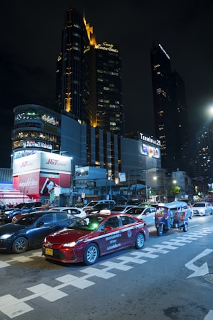 A bustling urban scene at night featuring a busy intersection with cars and a tuk-tuk. Skyscrapers with illuminated signs stand tall in the background, including buildings marked with 'Grande Centre Point' and 'Terminal 21'. Bright advertising billboards add vibrancy to the scene.