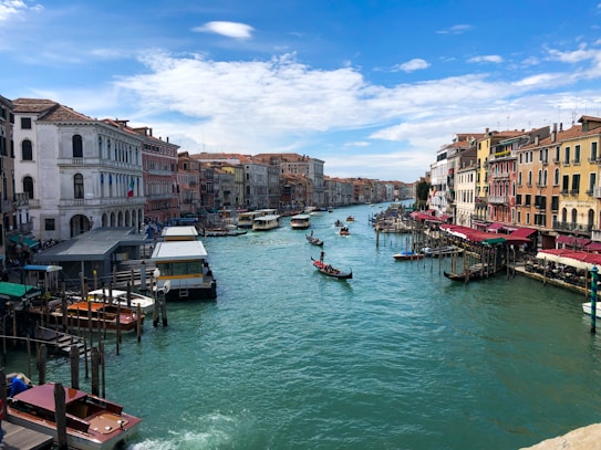 A picturesque canal lined with historic buildings featuring colorful fa&ccedil;ades. Several gondolas and boats are navigating the turquoise water. The sky is clear with a few clouds, creating a serene atmosphere.