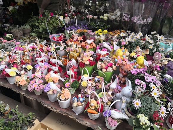 An assortment of small, decorative potted flower arrangements featuring a mix of colorful flowers including roses and daisies. The pots contain various decorative elements like artificial birds and small figurines. The scene is busy with a variety of colors and textures across the display.