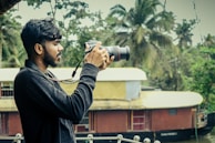 A person holding a DSLR camera is standing outdoors with a scenic background featuring lush green trees and a traditional houseboat on a river. The individual is focused on taking a photograph, demonstrating an interest in capturing nature.