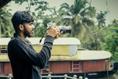 A person holding a DSLR camera is standing outdoors with a scenic background featuring lush green trees and a traditional houseboat on a river. The individual is focused on taking a photograph, demonstrating an interest in capturing nature.