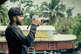 A person holding a DSLR camera is standing outdoors with a scenic background featuring lush green trees and a traditional houseboat on a river. The individual is focused on taking a photograph, demonstrating an interest in capturing nature.