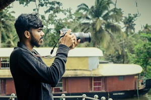 A person holding a DSLR camera is standing outdoors with a scenic background featuring lush green trees and a traditional houseboat on a river. The individual is focused on taking a photograph, demonstrating an interest in capturing nature.