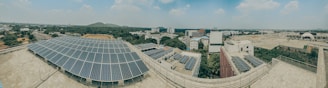 A panoramic view of an urban landscape with large solar panels prominently displayed on rooftops. The scene includes multiple industrial and residential buildings surrounded by lush greenery. The skyline has a few distant hills, and the sky is partially cloudy.