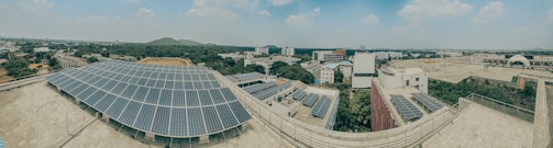 A panoramic view of an urban landscape with large solar panels prominently displayed on rooftops. The scene includes multiple industrial and residential buildings surrounded by lush greenery. The skyline has a few distant hills, and the sky is partially cloudy.