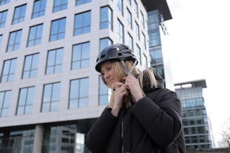 A person is fastening a strap on a black helmet while standing outdoors in front of a modern office building with numerous glass windows. The person is wearing a black jacket and appears to be focused on securing the helmet.