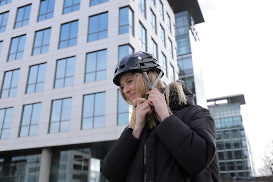 A person is fastening a strap on a black helmet while standing outdoors in front of a modern office building with numerous glass windows. The person is wearing a black jacket and appears to be focused on securing the helmet.