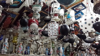 A crowded interior space filled with a variety of hanging lamps and chandeliers, featuring ornate designs and a mix of colors and materials. The walls are adorned with framed paintings. The collection includes vases and other decorative pieces arranged closely together.