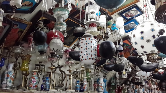 A crowded interior space filled with a variety of hanging lamps and chandeliers, featuring ornate designs and a mix of colors and materials. The walls are adorned with framed paintings. The collection includes vases and other decorative pieces arranged closely together.