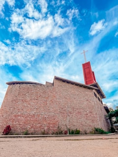 A large brick building with a distinct architectural design features a red tower topped with a cross, set against a vibrant blue sky with scattered clouds. Surrounding the structure, some greenery is visible at the base.