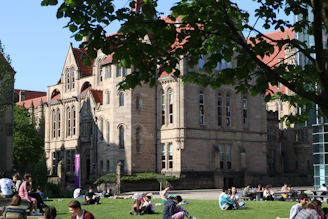 Historic UK university building with students sitting on the lawn during a sunny day.