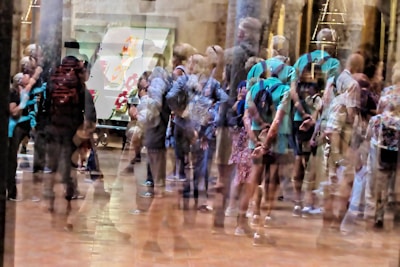 A blurred image with multiple overlapping impressions of people standing in a gallery space. The scene is bustling with movement and activity, suggesting a crowded environment. The walls have artworks displayed and there are columns visible in the background.