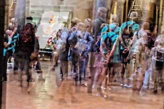 A blurred image with multiple overlapping impressions of people standing in a gallery space. The scene is bustling with movement and activity, suggesting a crowded environment. The walls have artworks displayed and there are columns visible in the background.