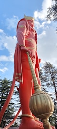 A large statue of a mythological figure with pinkish-red clothing and a golden crown, holding a mace. The statue is surrounded by trees under a blue sky with scattered clouds.