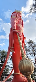 A large statue of a mythological figure with pinkish-red clothing and a golden crown, holding a mace. The statue is surrounded by trees under a blue sky with scattered clouds.